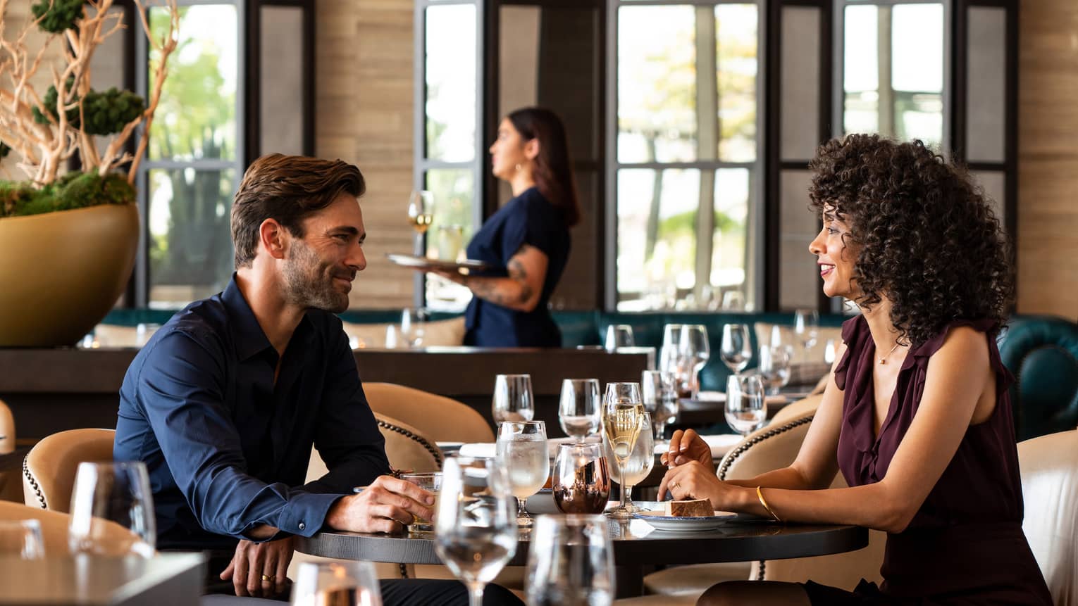 A man and woman enjoy a date at trendy cinderhouse – furnished with black square tables, camel leather colored chairs, and modern sconce lighting 