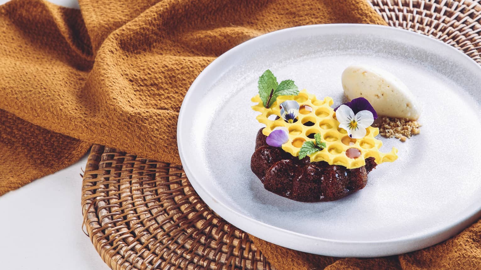 A rich chocolate mini-cake topped with honeycomb-shaped white chocolate garnished with mint leaves and dainty flower petals