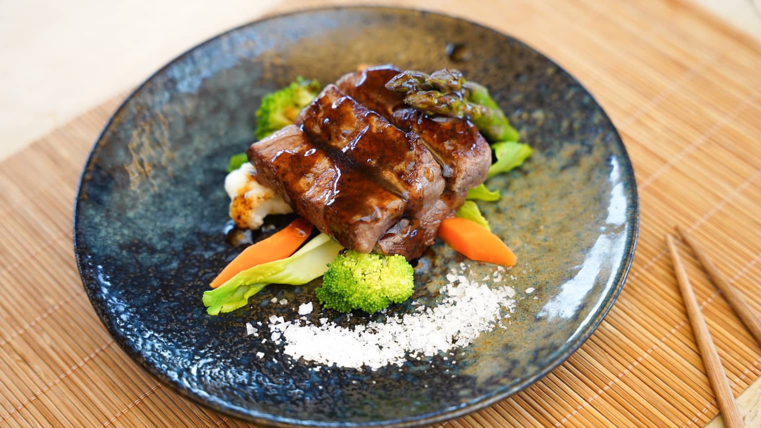 Beef tenderloin with sauce atop mixed vegetables on blue-grey glazed ceramic plate with chopsticks nearby