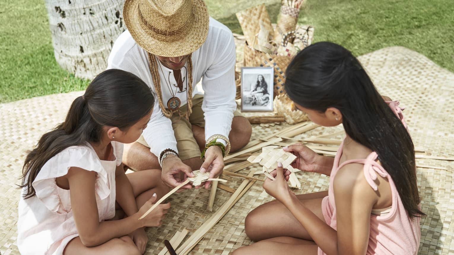 Adult helps two young girls learn how to weave as they sit on a woven blanket