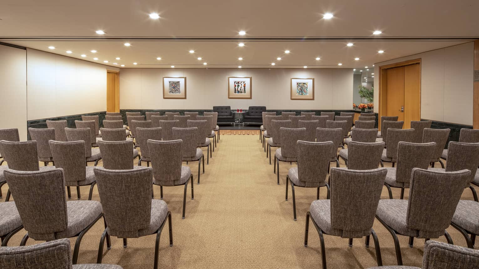 Rows of gray chairs face the front of a function room with tan walls and a few gallery frames 