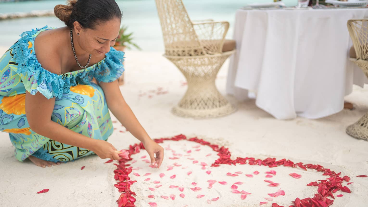 Woman arranges red rose petals into heart shape on sand beach