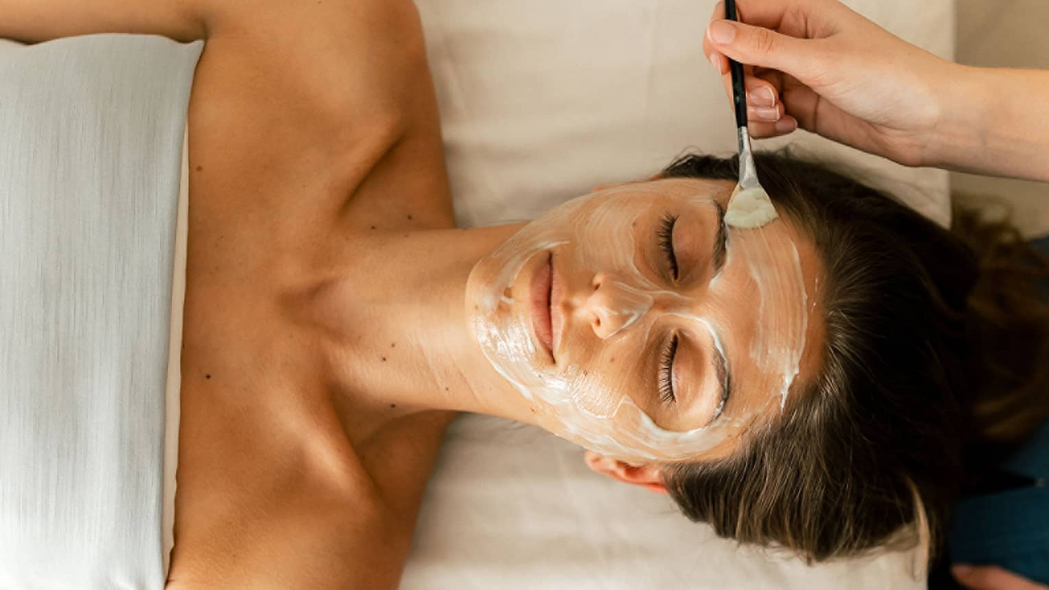 A person holding a small brush applies a white face mask treatment onto a woman's face