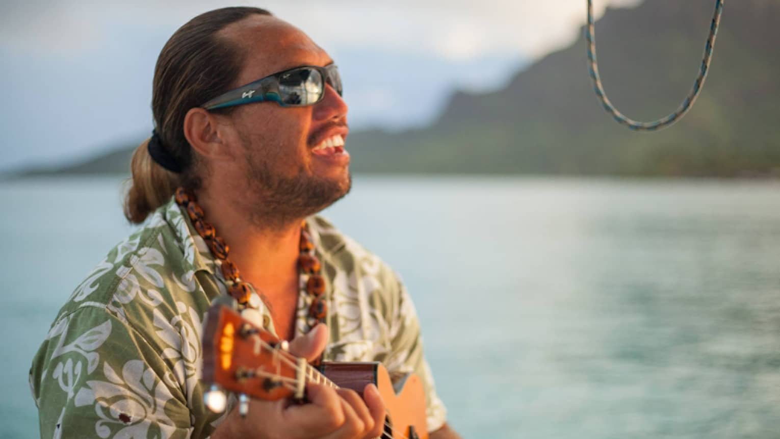A musician wearing sunglasses and beads plays the ukulele beside the ocean, face aglow from the sunset.