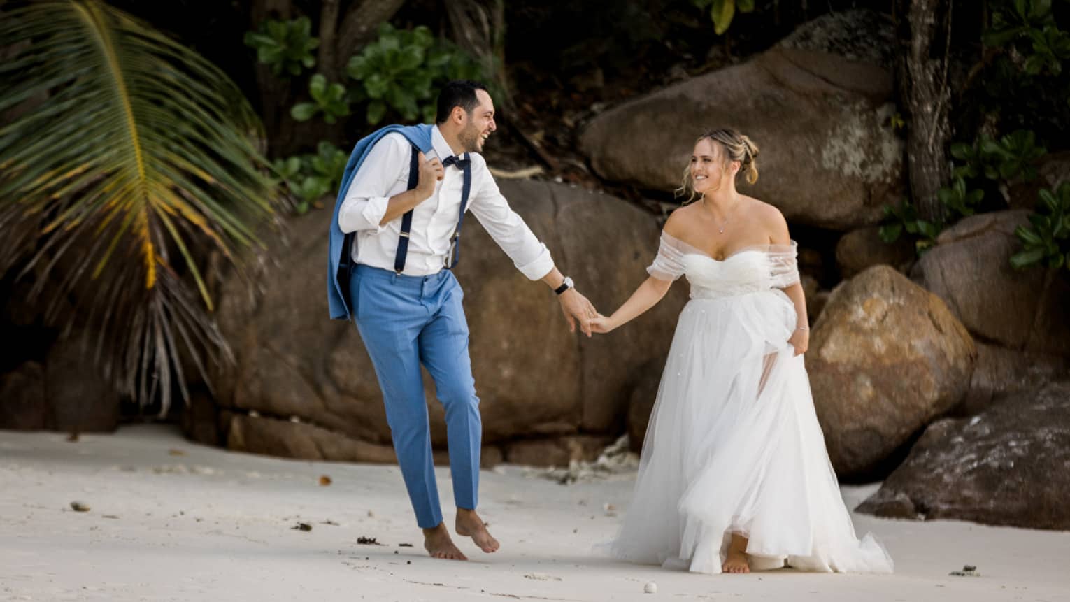 Groom in blue suit holding bride's hand as they stand on beach