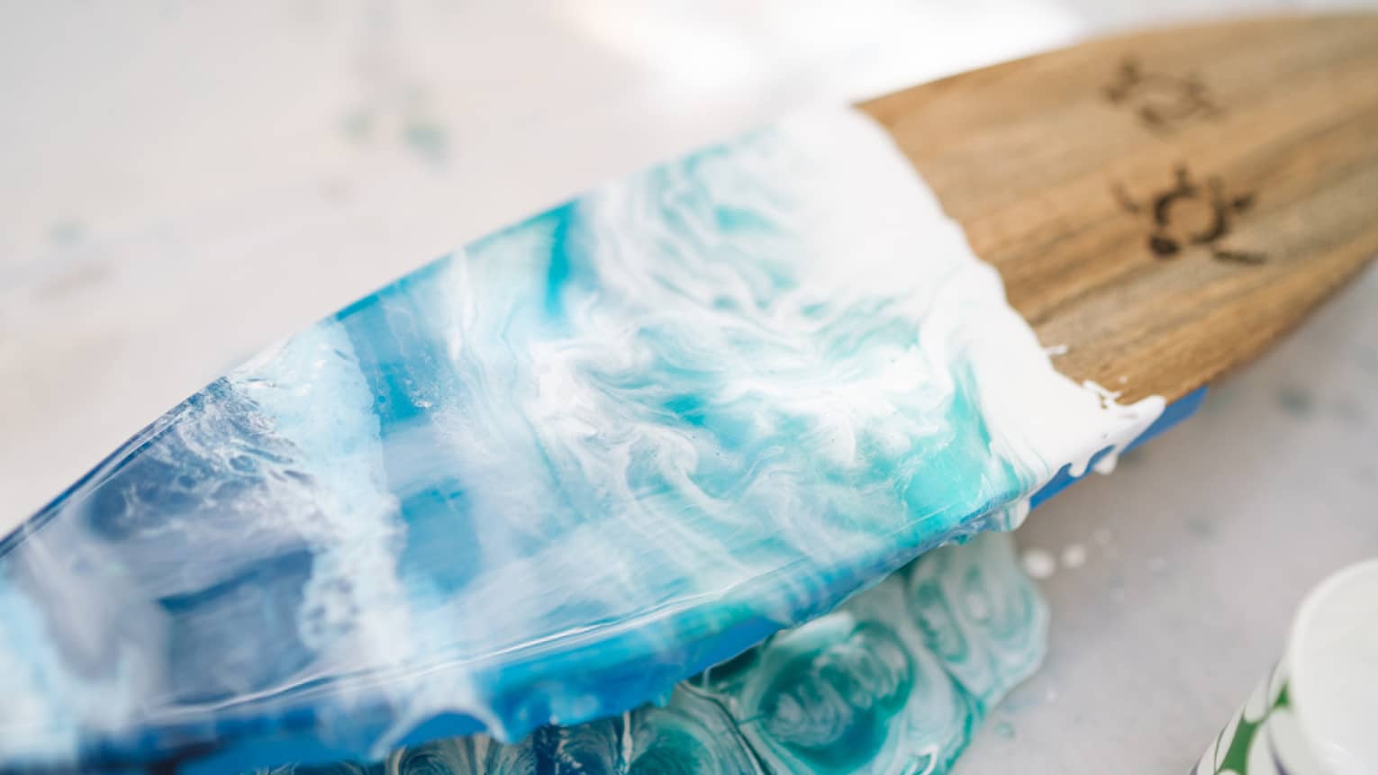 Close-up of wooden surfboard half-painted in deep to turquoise blues with white wave-like swirls and turtle outline drawings.