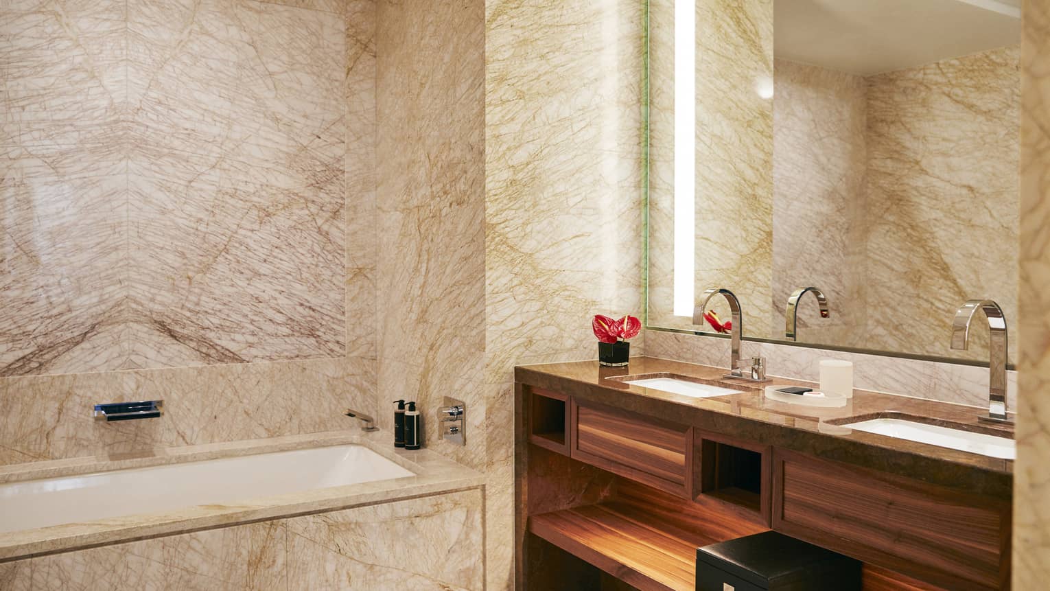 Luxury hotel bathroom with marble walls and surfaces, bathtub, double sinks with modern fixtures and a softly-lit mirror above a polished wooden vanity