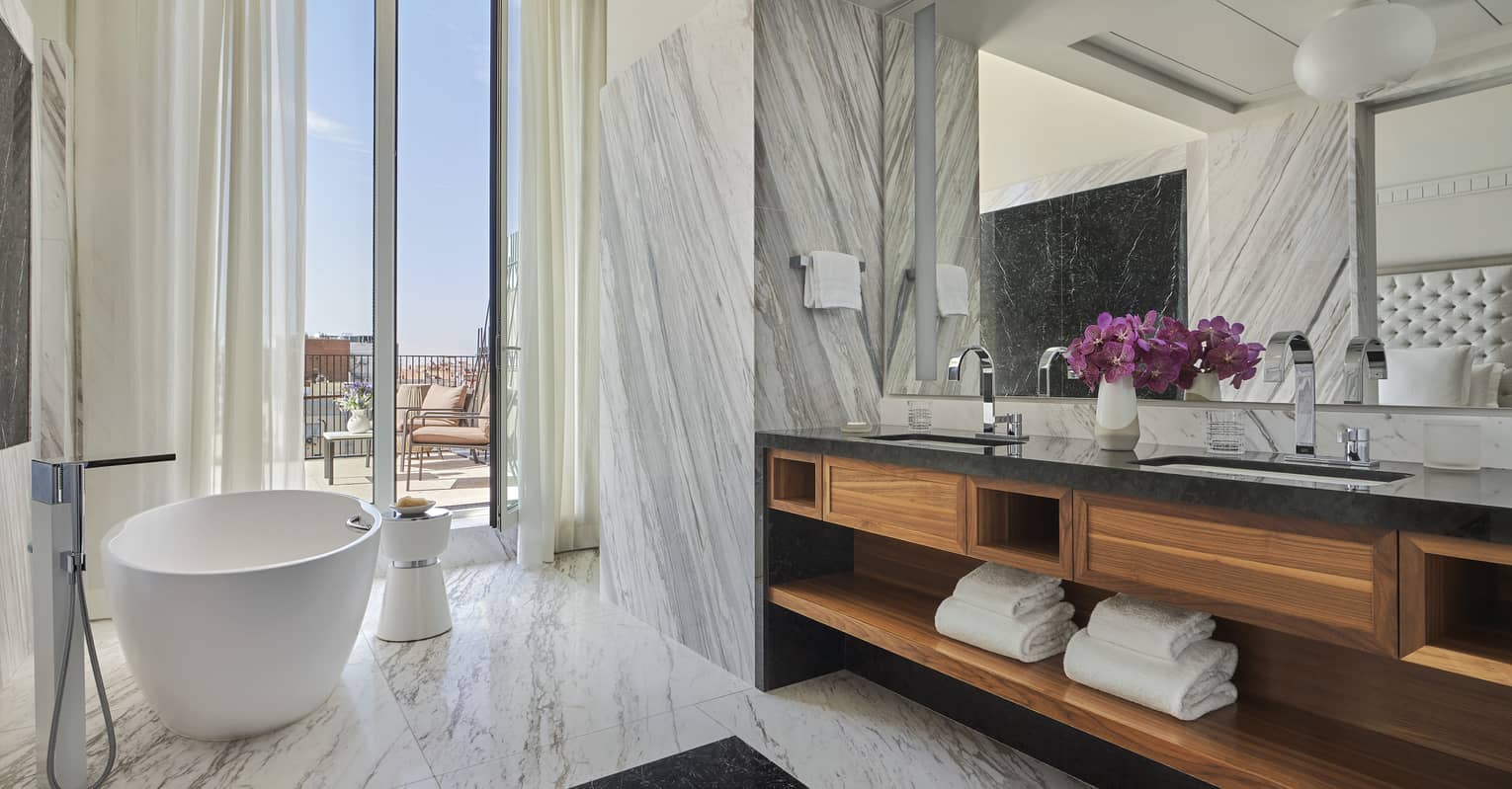 Luxury hotel bathroom with a freestanding tub, dual-sink vanity with wood accents, large mirror, marble walls and access to a private terrace