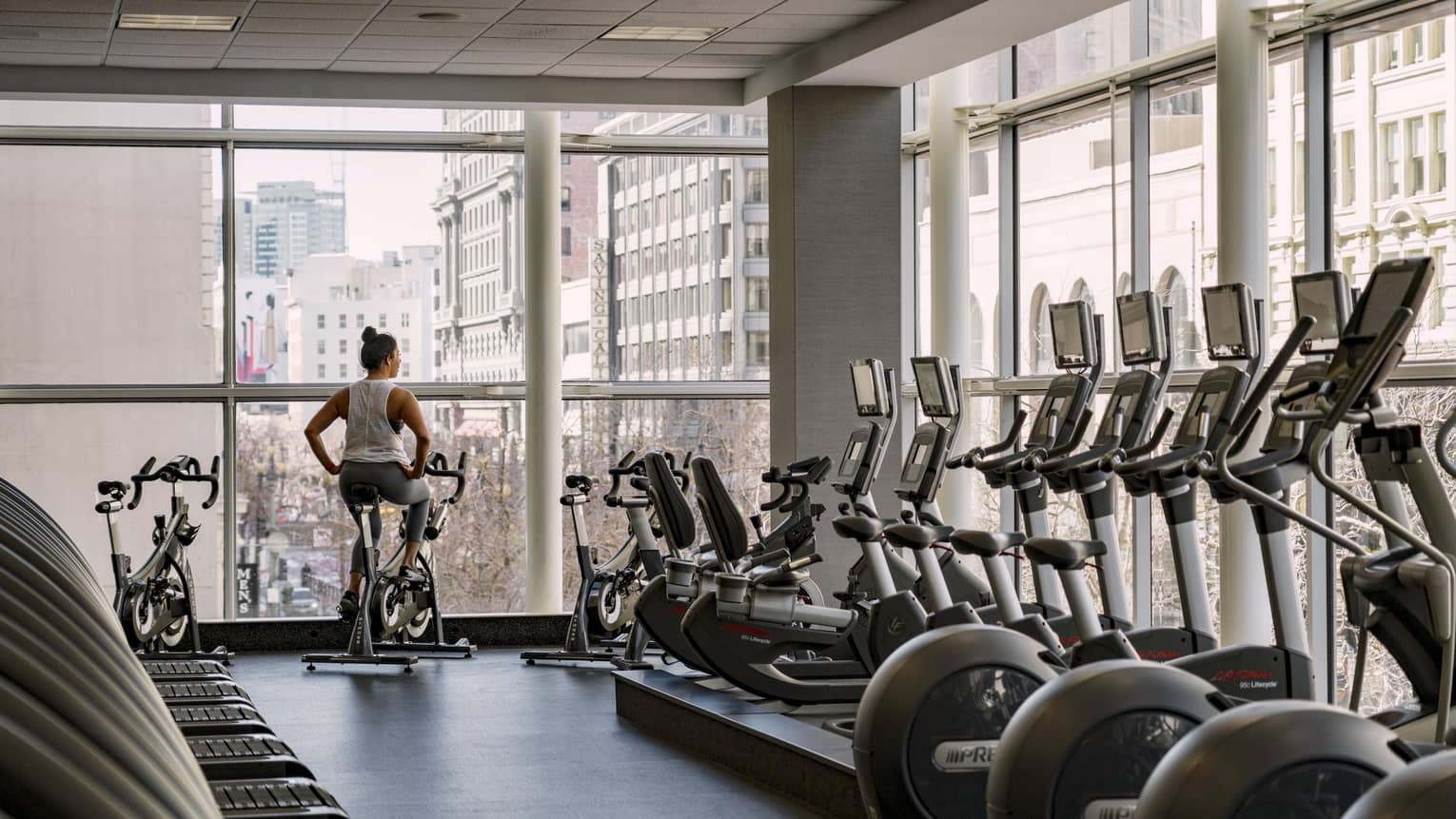 A woman rides a stationary bike in a hotel fitness center with floor-to-ceiling windows overlooking a city view