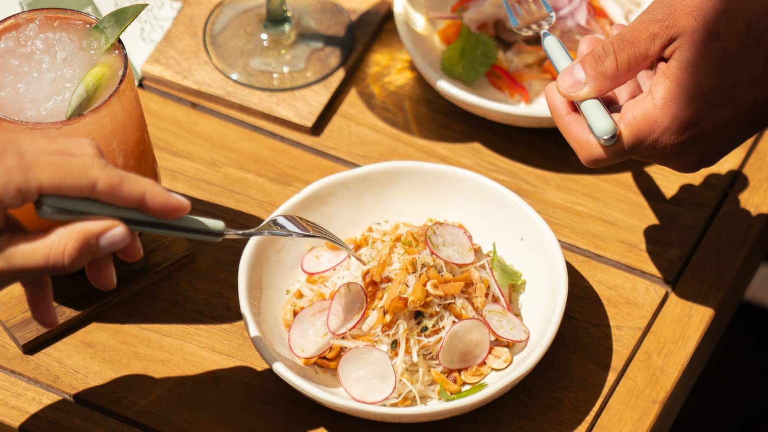 A wood table with small dishes of food items and cocktails in stemware and a tumbler. Two people reach for bites of food with forks.