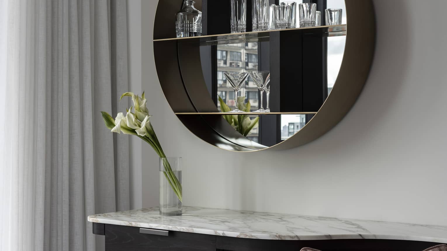 A modern vanity contains a marble countertop, round mirrored shelf displaying glassware and pink upholstered stool. A vase with white flowers sits on the vanity, complemented by soft lighting.