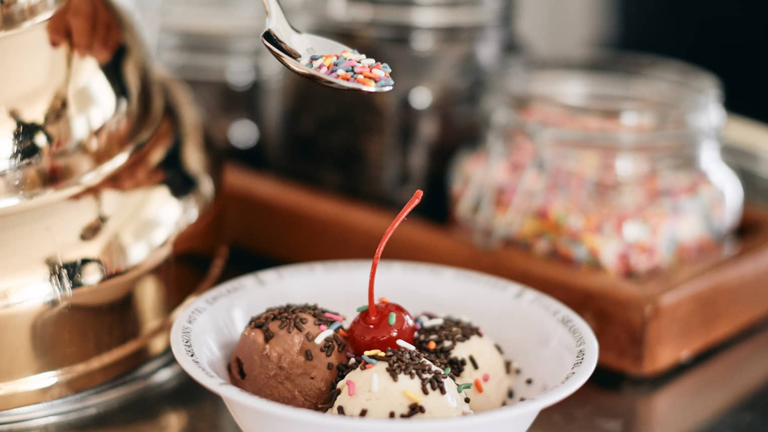 A member of staff creating a colorful ice cream bowl for a guest, there are various flavours in the bowl, sprinkles, and a cherry.