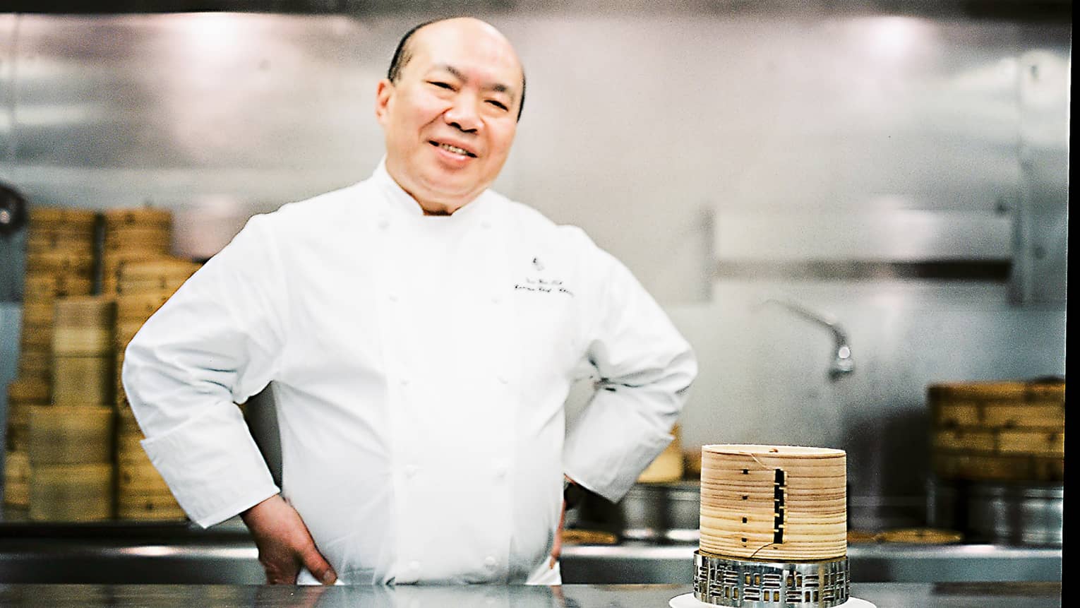 Chef Chan Yan Tak stands smiling in a professional kitchen with his hands on his hips, wearing a white uniform. Bamboo steamers are on the counter. 