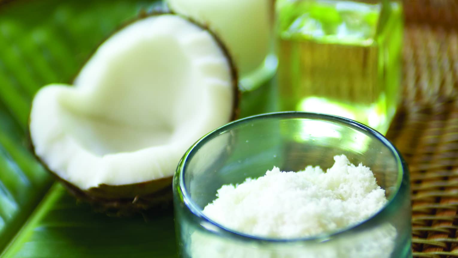 Half of a cracked coconut shell next to small glass bowl with coconut flakes