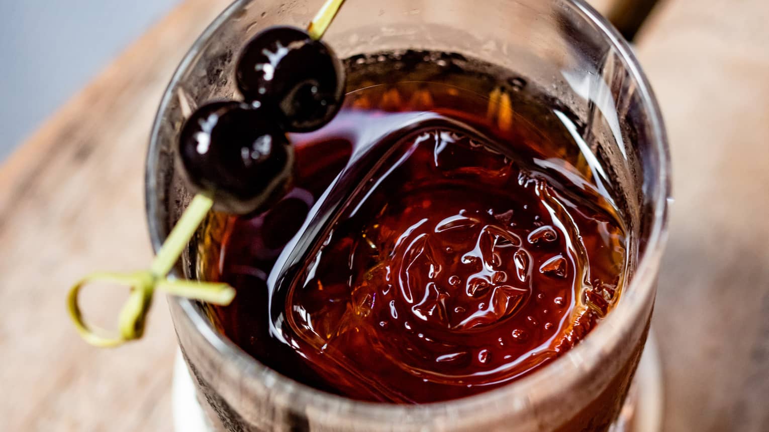 A dark coloured cocktail with two cherries on top of the glass.