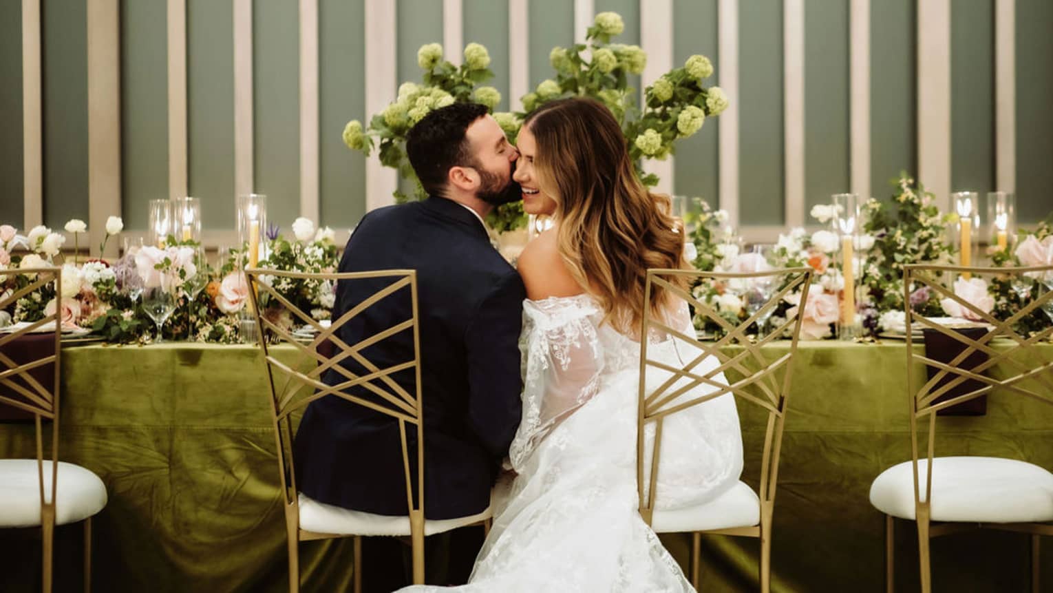Man in tux and woman in wedding dress sitting together at a table with a bouquets of flowers on it.