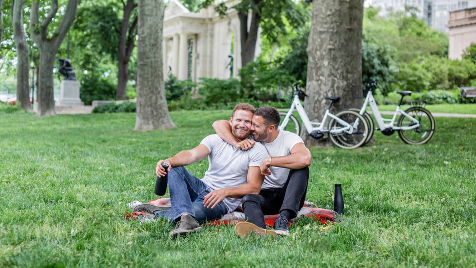 Tow men hugging and sitting on a picnic blanket near trees.