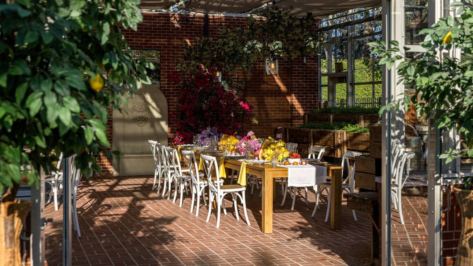 Wooden rectangular table topped with flowers and flanked by eight white chairs sits in a covered patio surrounded by windows and greenery