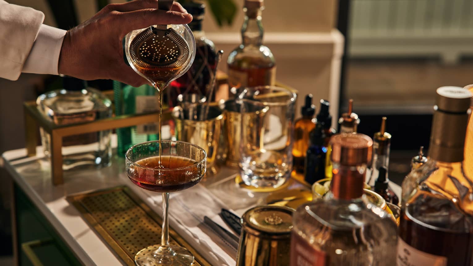A bartender pouring a drink into a cocktail glass with bottles of liquor around.