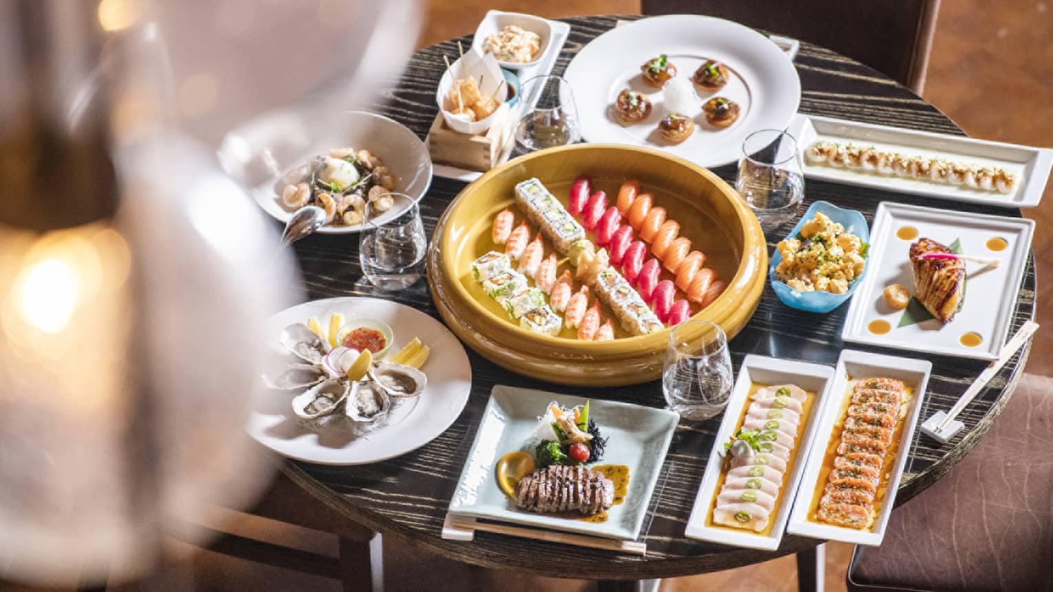 Small round table filled to the brim with a variety of square and round dishes filled with food, all surrounding a round gold platter with five rows of sushi in the center
