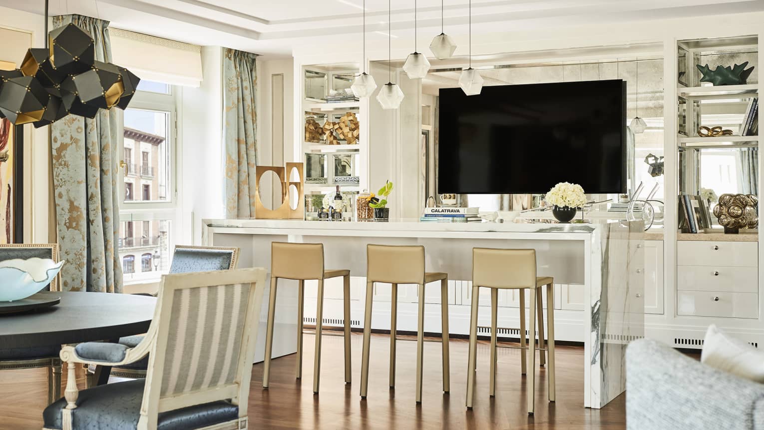 Luxury hotel suite lounge area with a marble counter, beige barstools, mounted TV, decorative shelving, dining table and large windows with patterned curtains
