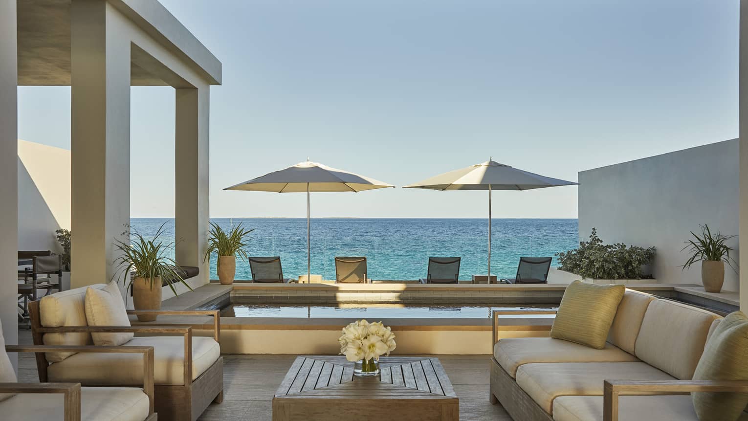 Outdoor terrace with cushioned seating, a wooden table, two umbrellas and ocean views beyond a private pool