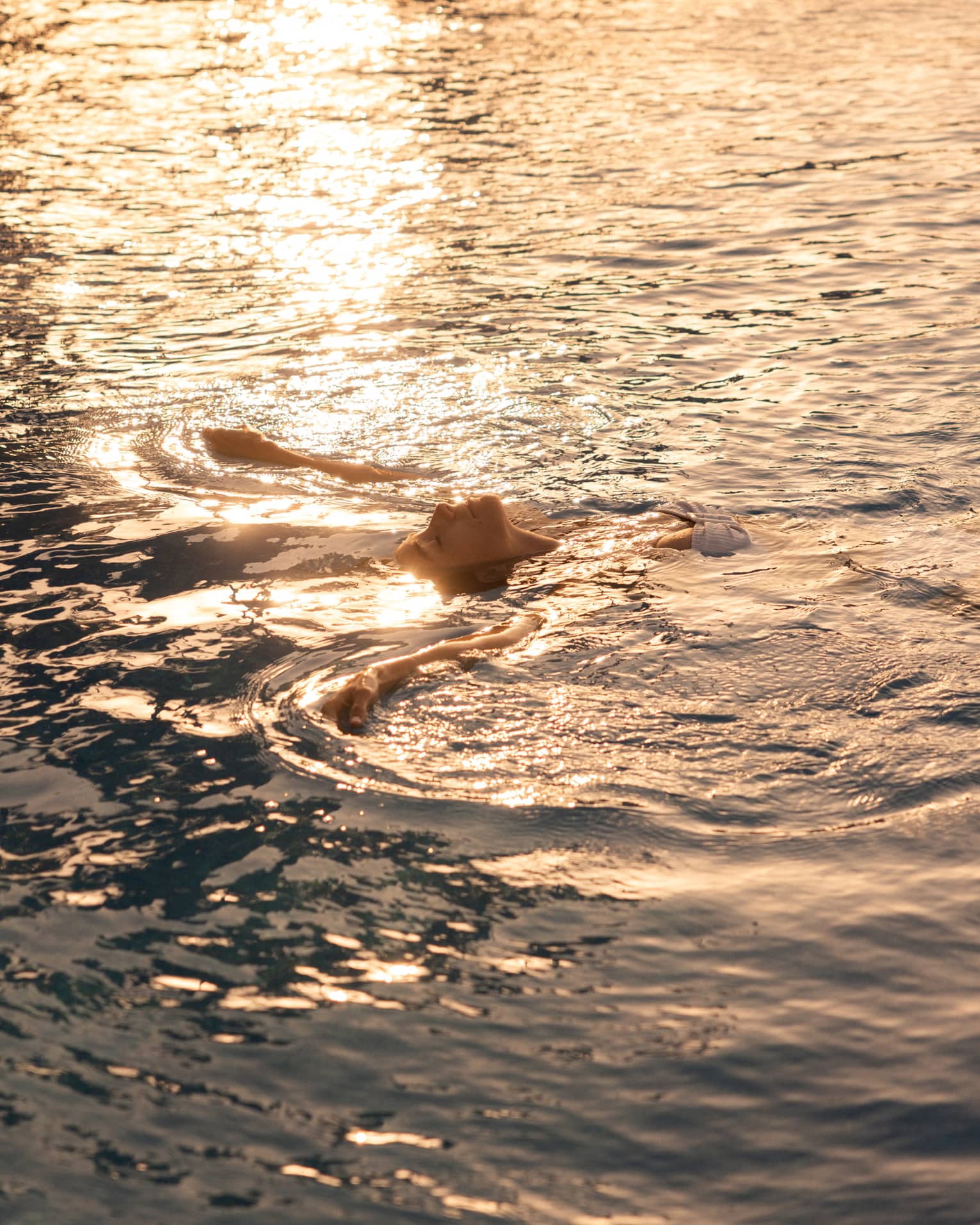 A person floating on their back in the ocean, with sunlight rippling across the water.