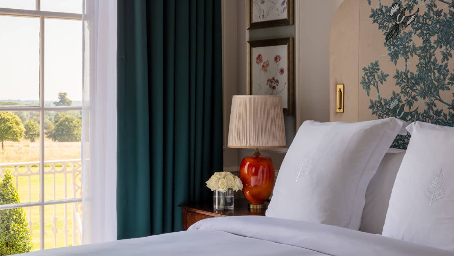 Guest room with bed with white linens and embroidered headboard, cherry bed side table and red lamp, window with blue drapes overlooking the garden