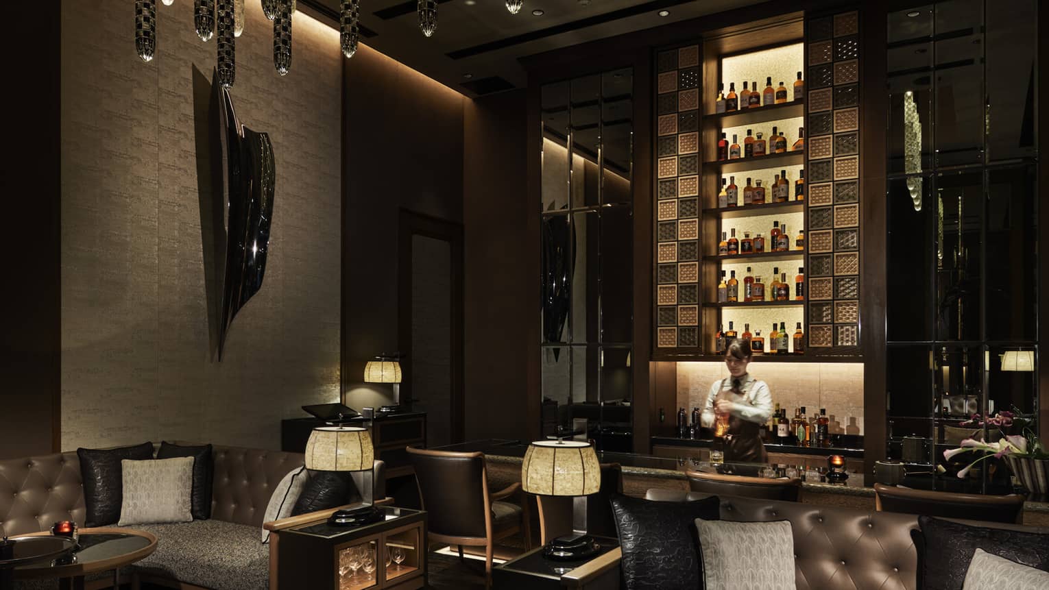 Bar area with leather loveseats, low lighting, fully stocked bar wall, bartender mixing drink behind bar