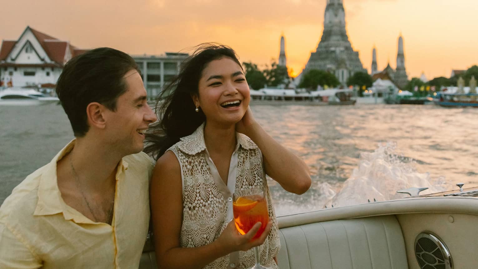 Smiling couple sits at back of boat holding cocktails on Chao Phraya River sunset tour