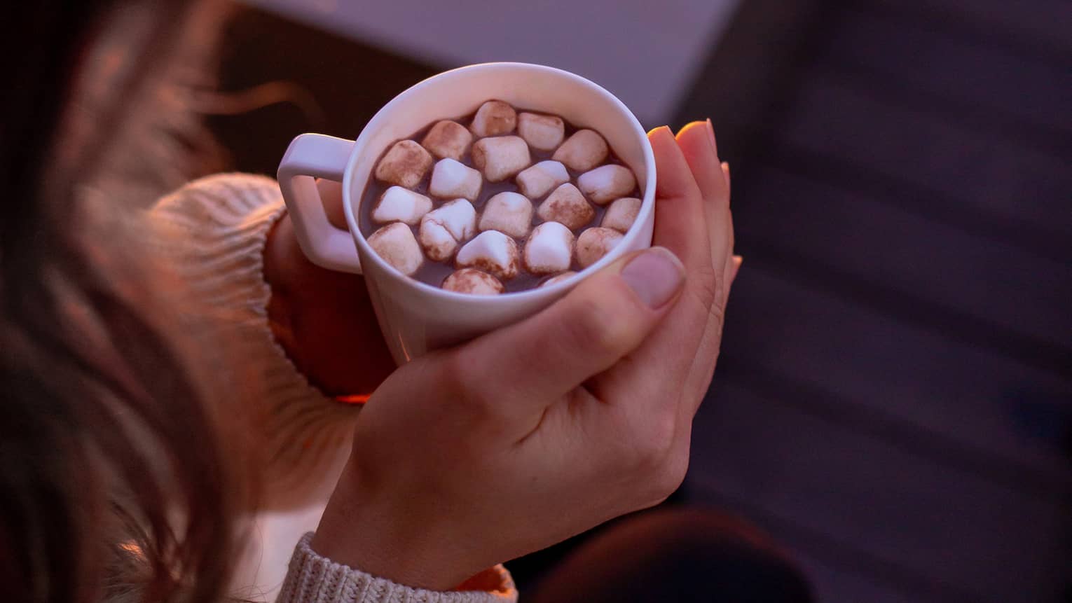 A woman holding hot chocolate by a fire.