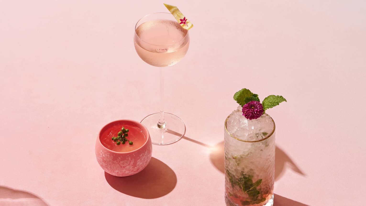 Three cocktails, each in a different type of glass, sit on a pink gradient backdrop
