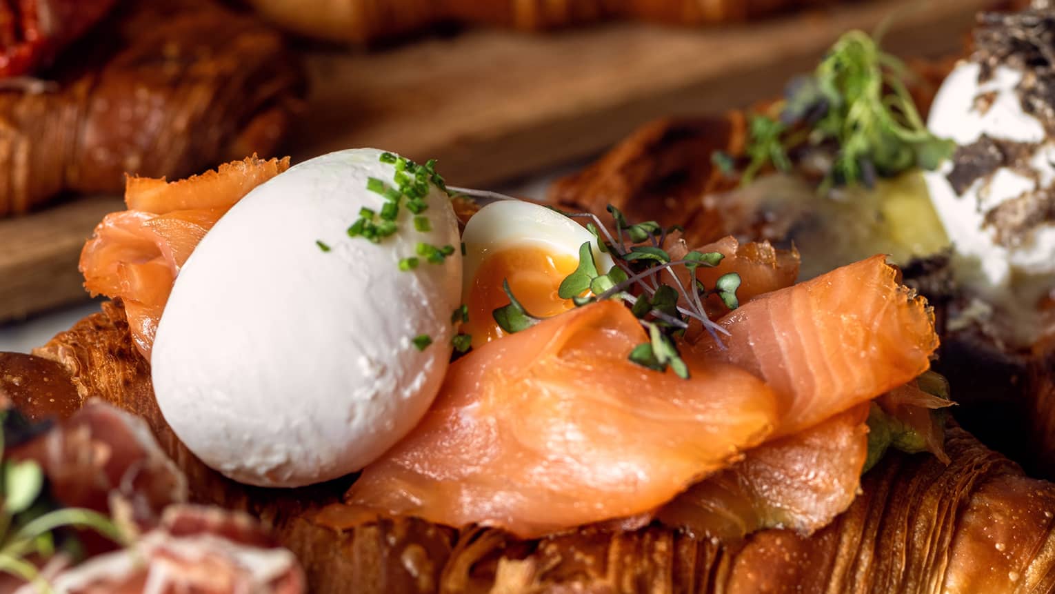 Croissants topped with smoked salmon, whole poached eggs and fresh herbs on boards