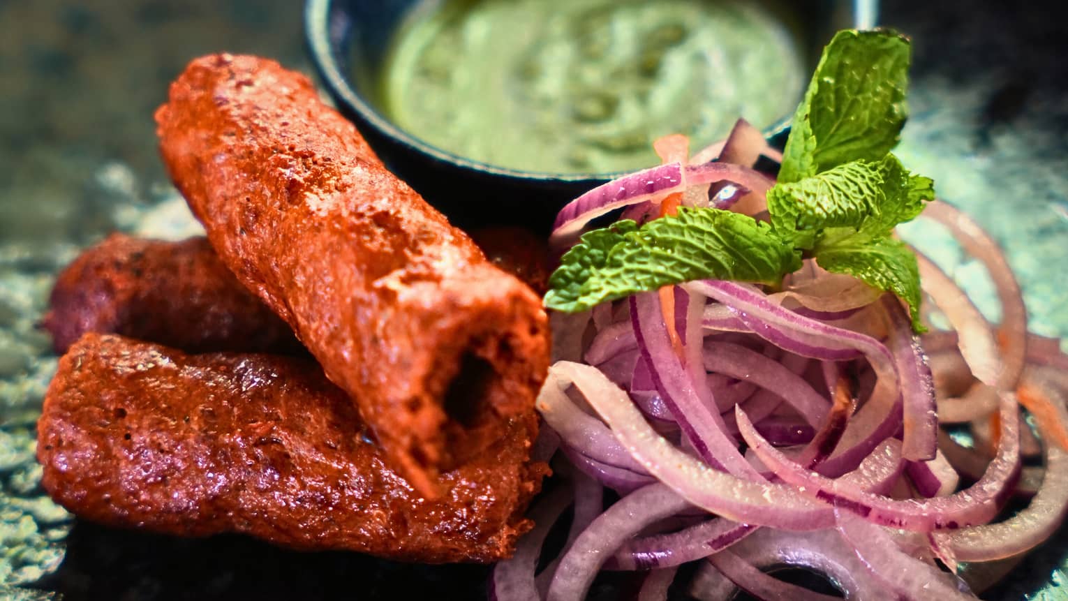 Food items with garnishes and a small bowl of sauce, all together on a dark-glazed plate
