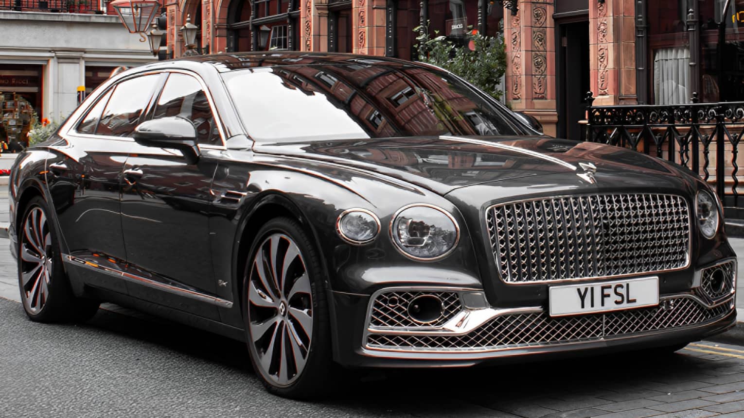 Close-up front view of shiny black modern Bentley with an elaborate grille parked in front of a restored Victorian building.