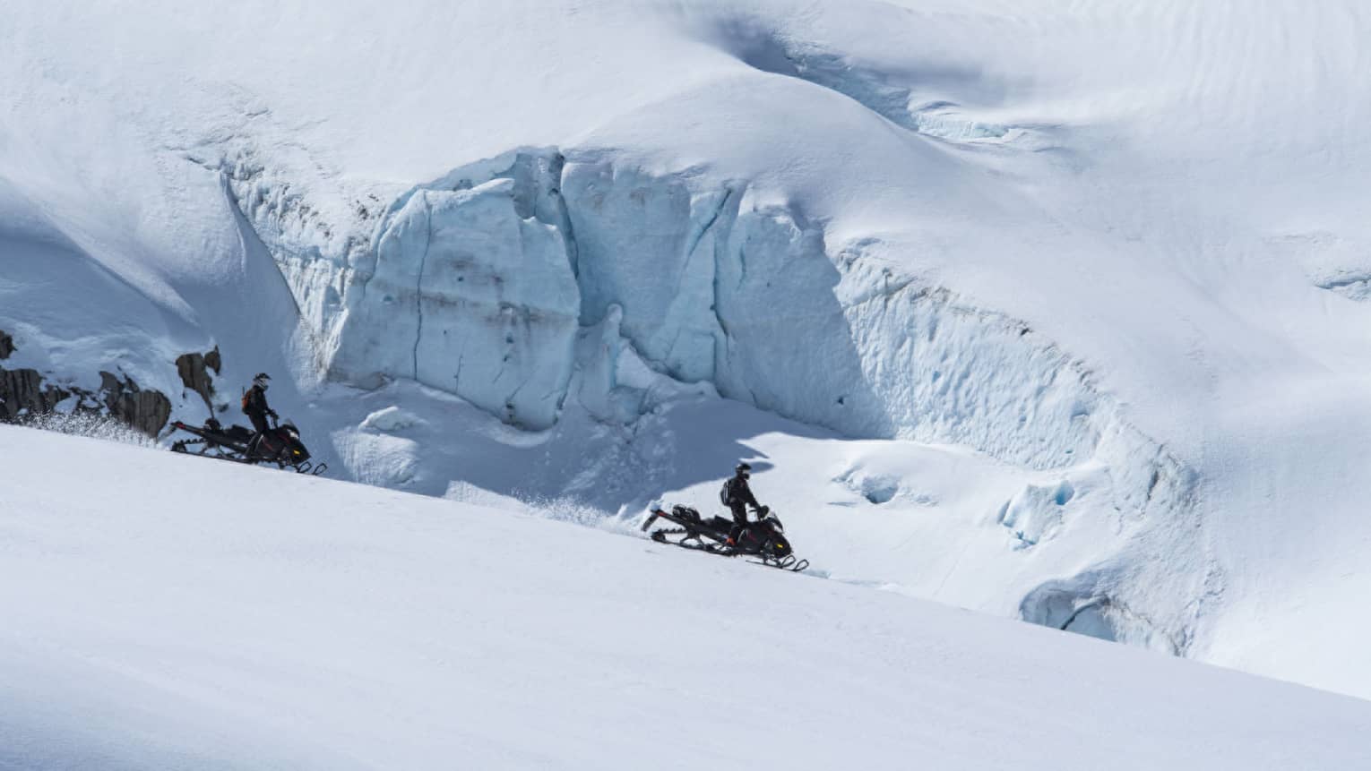 Side view of two distant black-clad snowmobilers coasting down a white slope alongside an enormous wall of ice and snow.