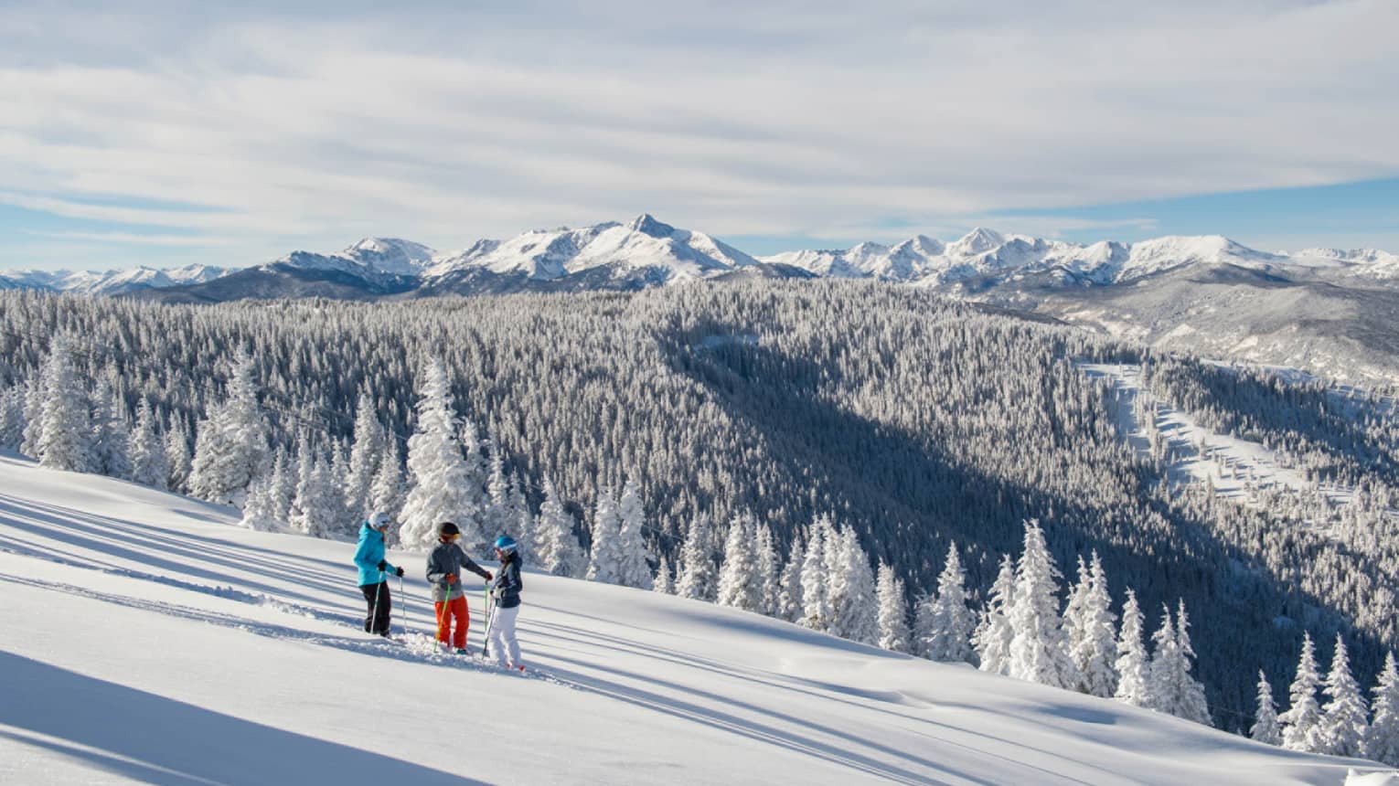 Three guests on skis pause on the ski hill, snow capped trees and mountains in distance 