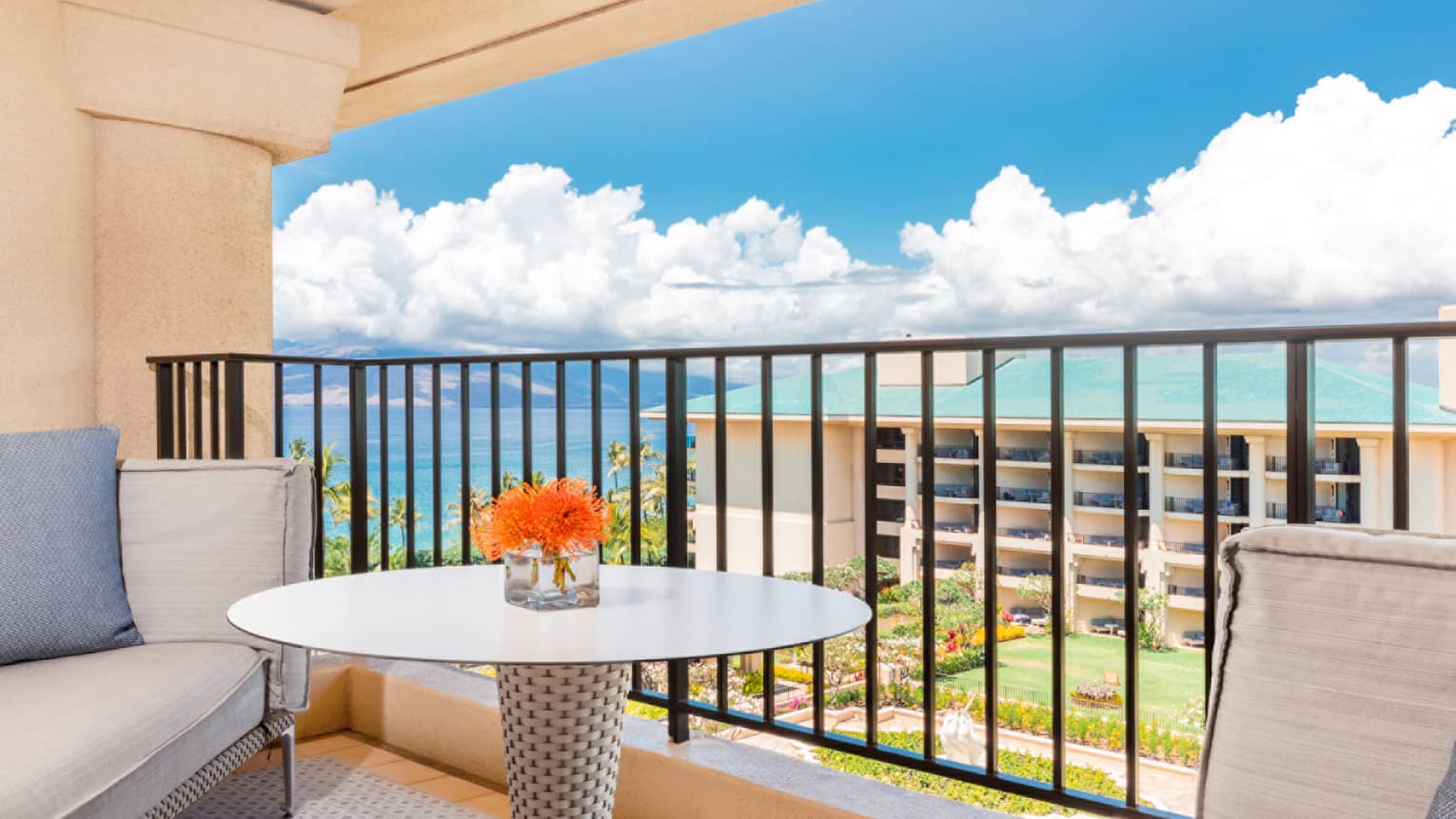 Resort balcony with round table, orange flowers, cushioned seating and ocean view under a bright blue sky