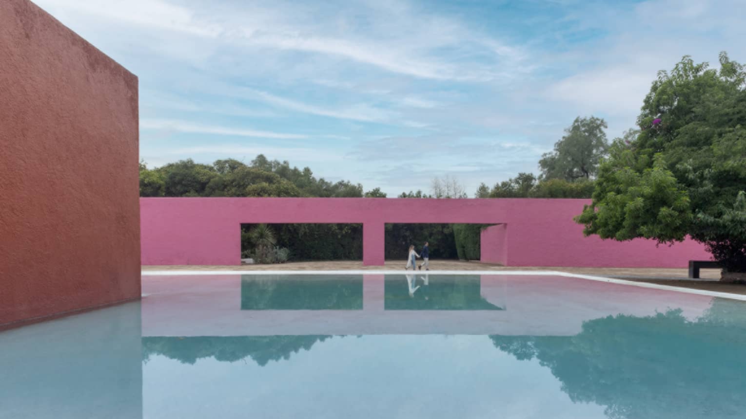 Two people standing next to a large pool with a pink wall behind them, trees can be seen through two large holes in the pink wall.