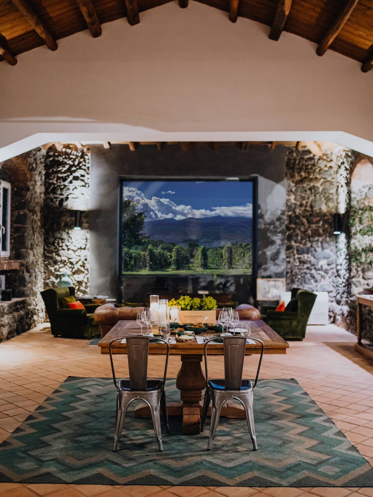 A wooden dining table set with floral arrangements and glassware, in a room with a window overlooking mountains and a vineyard