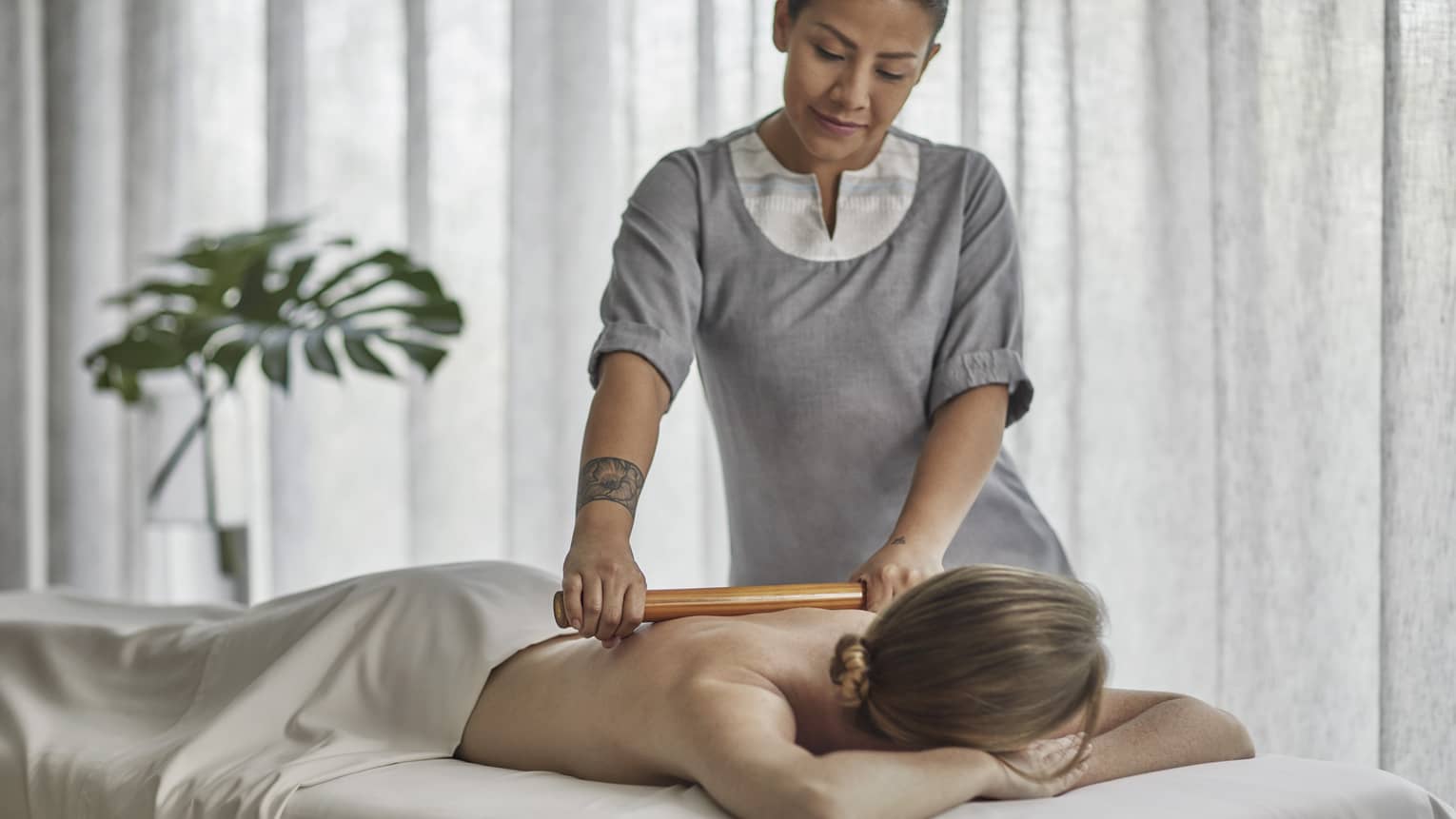 Spa therapist wearing a grey tunic rolls a wooden roller on the bare back of a guest who is laying on a massage table 