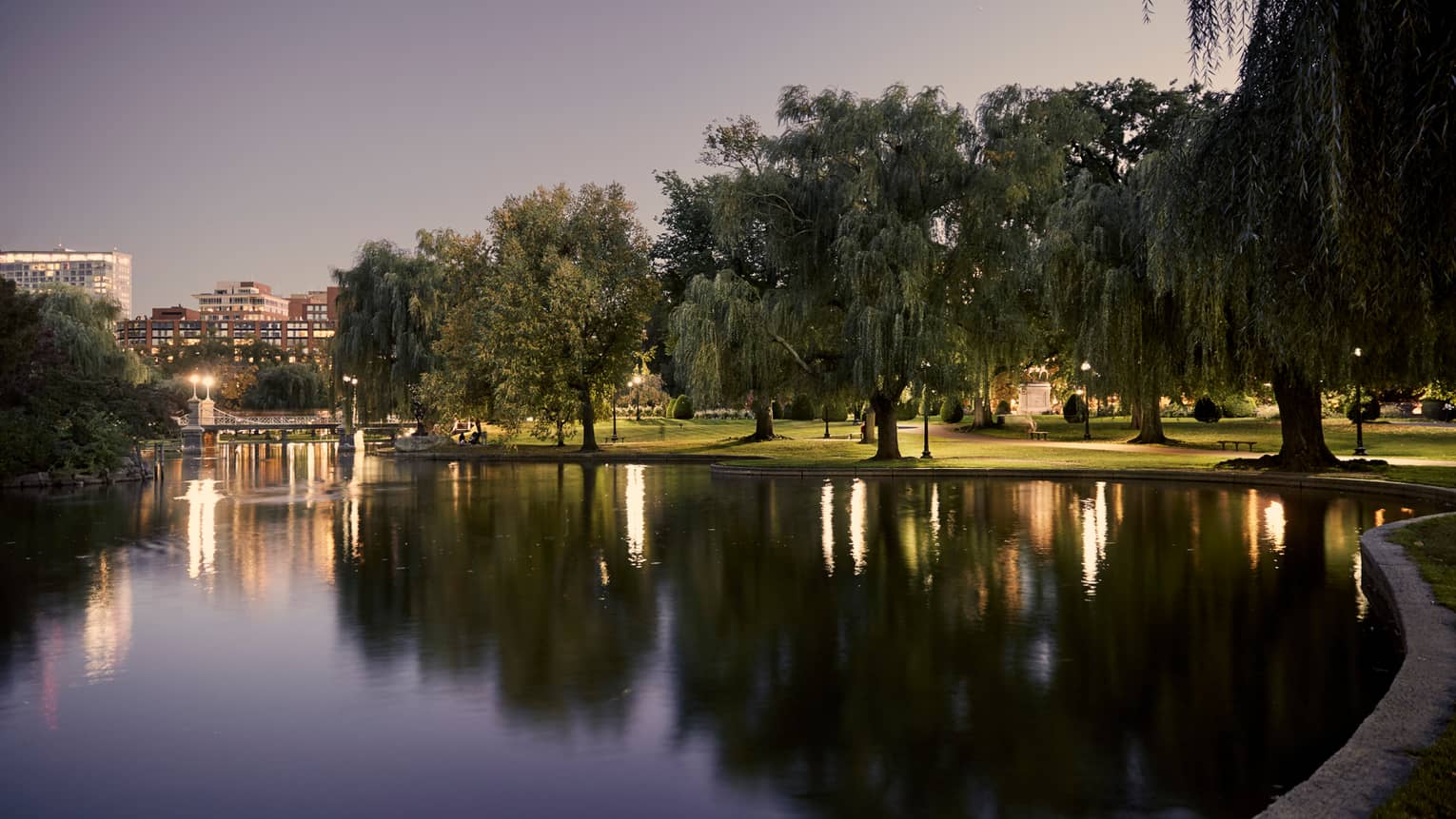Boston Public Garden pond lined with large trees, lights at dusk