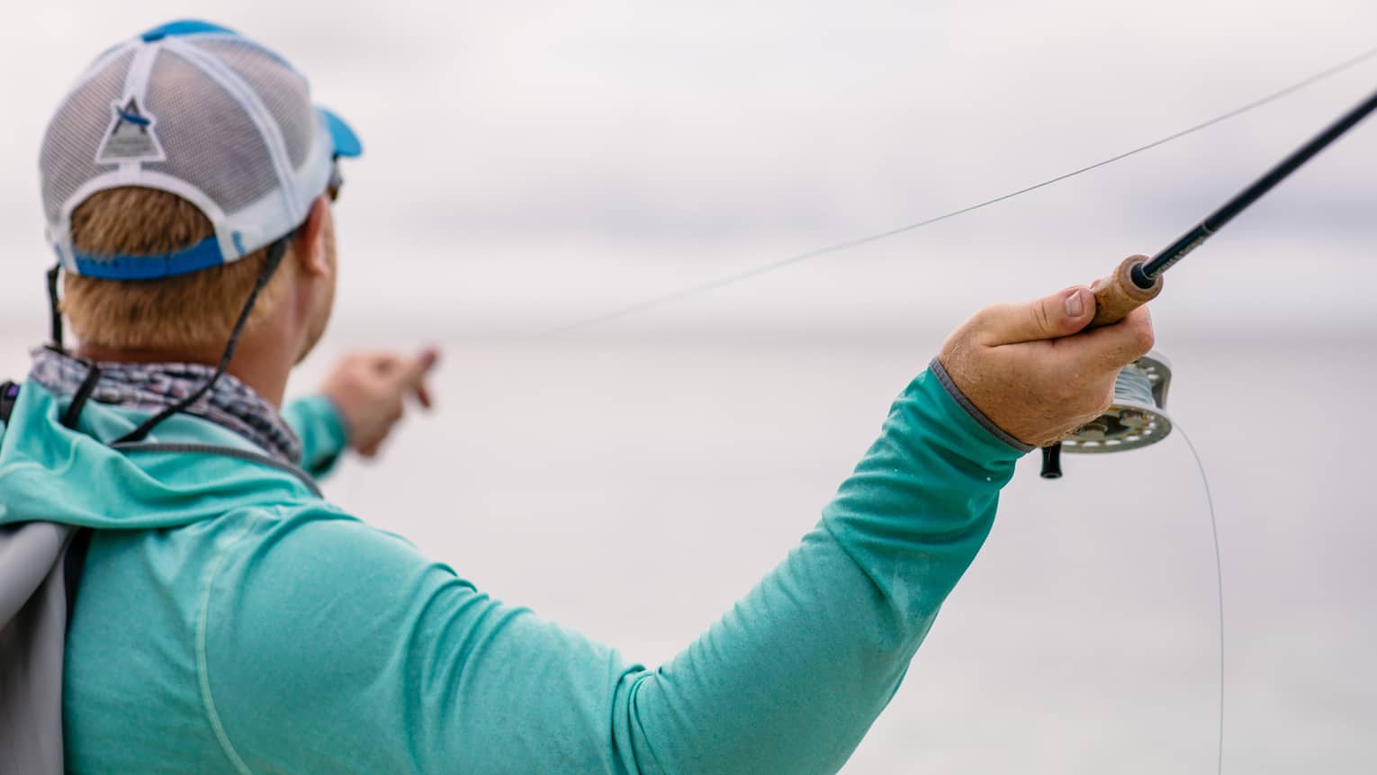 A fisherman facing away holds his fishing line in his left hand and draws back his rod with his right, readying to cast. 