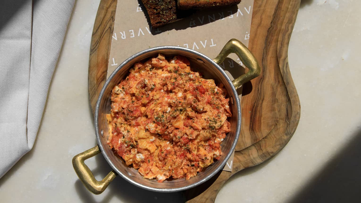 Pan of scrambled eggs with fresh tomato, feta cheese, oregano on cutting board with side of bread