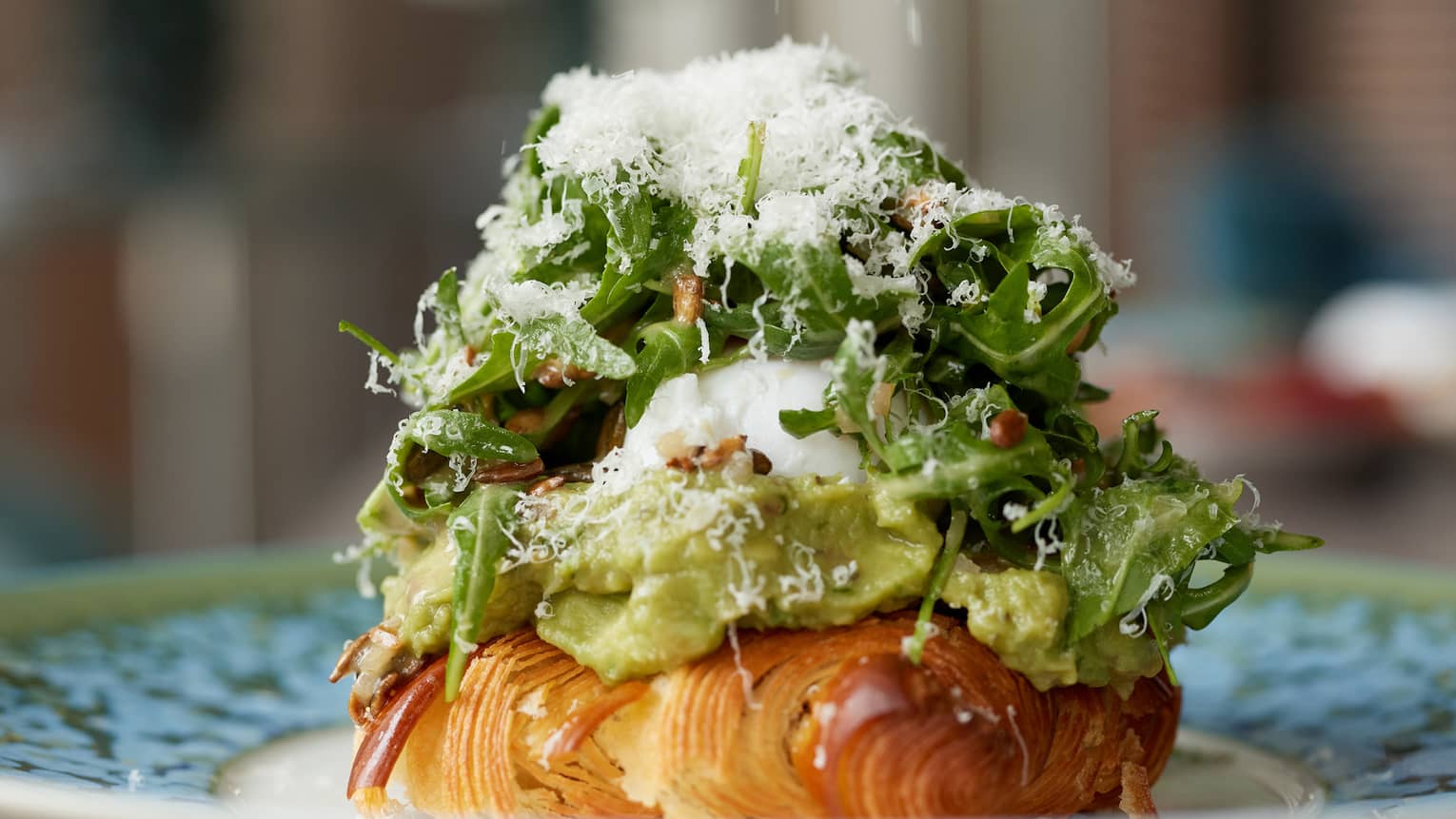 A close-up of a plate holding a croissant covered with avocado, egg and arugula salad topped with grated parmesan. 