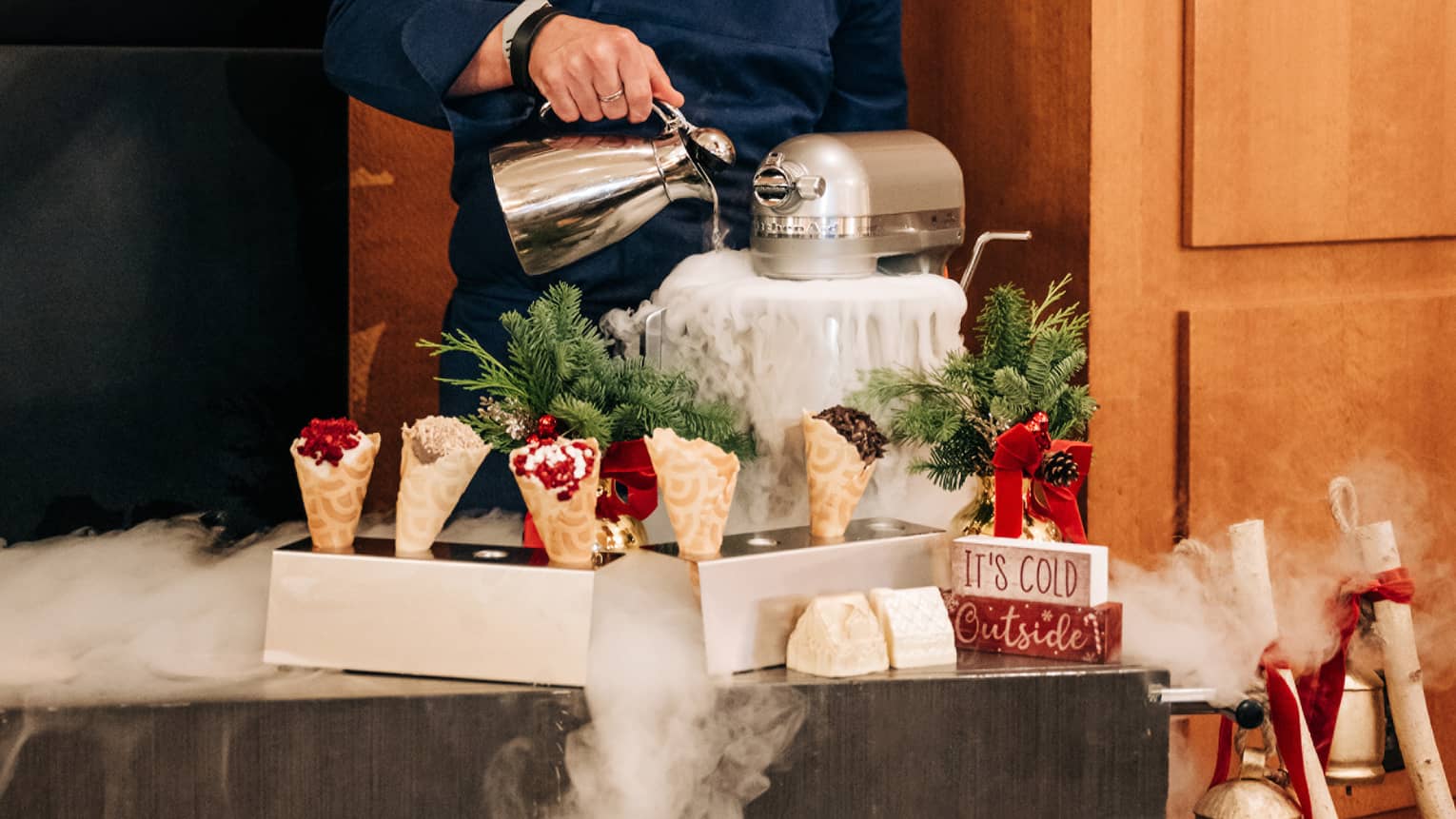 Chef pours liquid nitrogen into a mixer in front of a cart topped with waffle cones and holiday decor