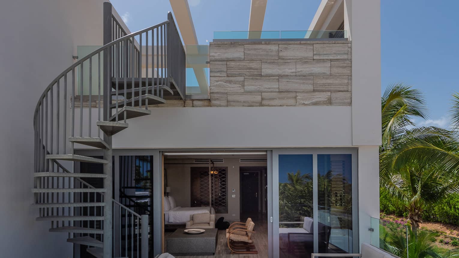 Luxury rooftop suite with sliding glass doors, a private balcony with seating, a spiral staircase and lush greenery in the background