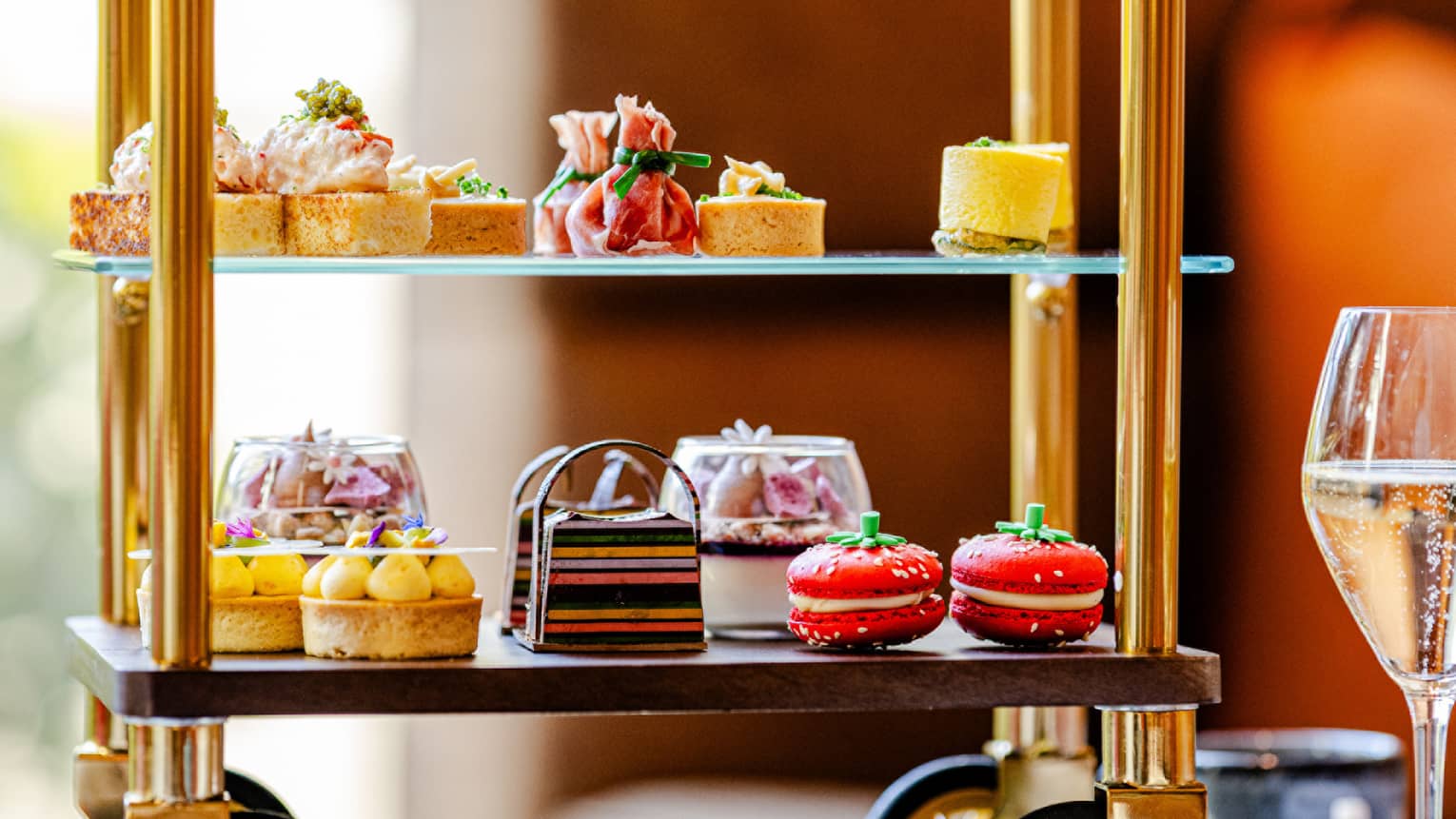 A tiered serving tray on small wheels holds an assortment of colourful small bites. It sits on a tabletop next to a stemmed glass, a teapot, and a teacup and saucer.
