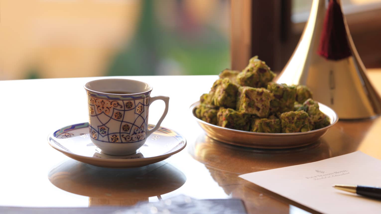 Turkish coffee in decorative cup beside plate with square treats, pen and notepad