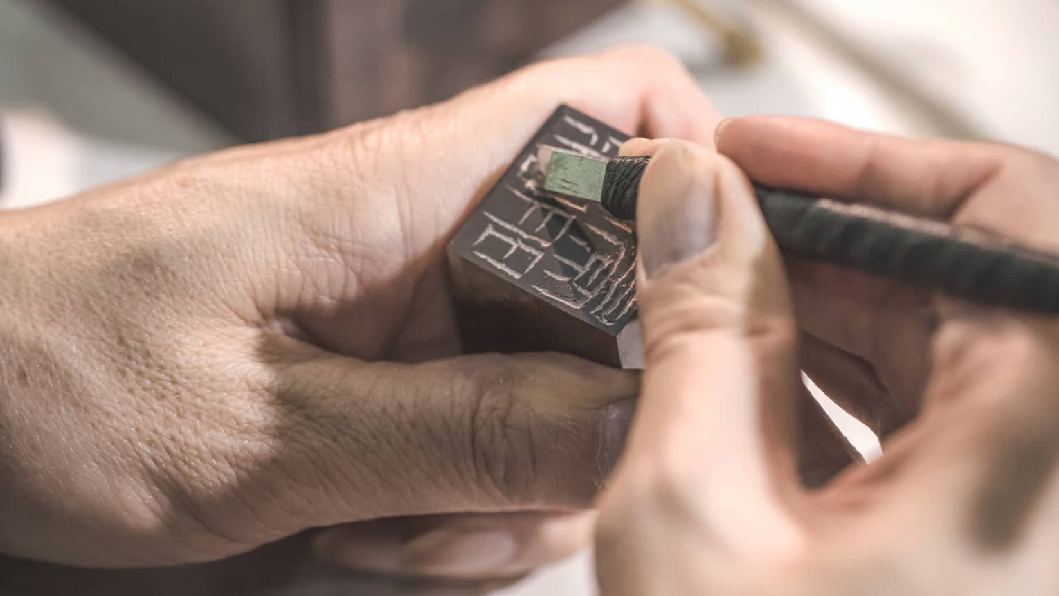 A hand holds a small block of dark stone while another hand carefully uses a tiny chisel to carve symbols into its surface.